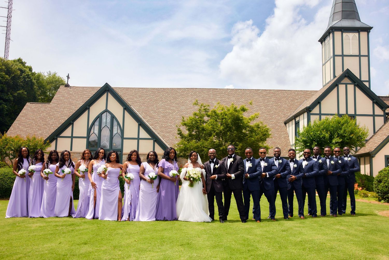 A wedding party lineup poses outdoors in front of a church with a tall spire on a sunny day.