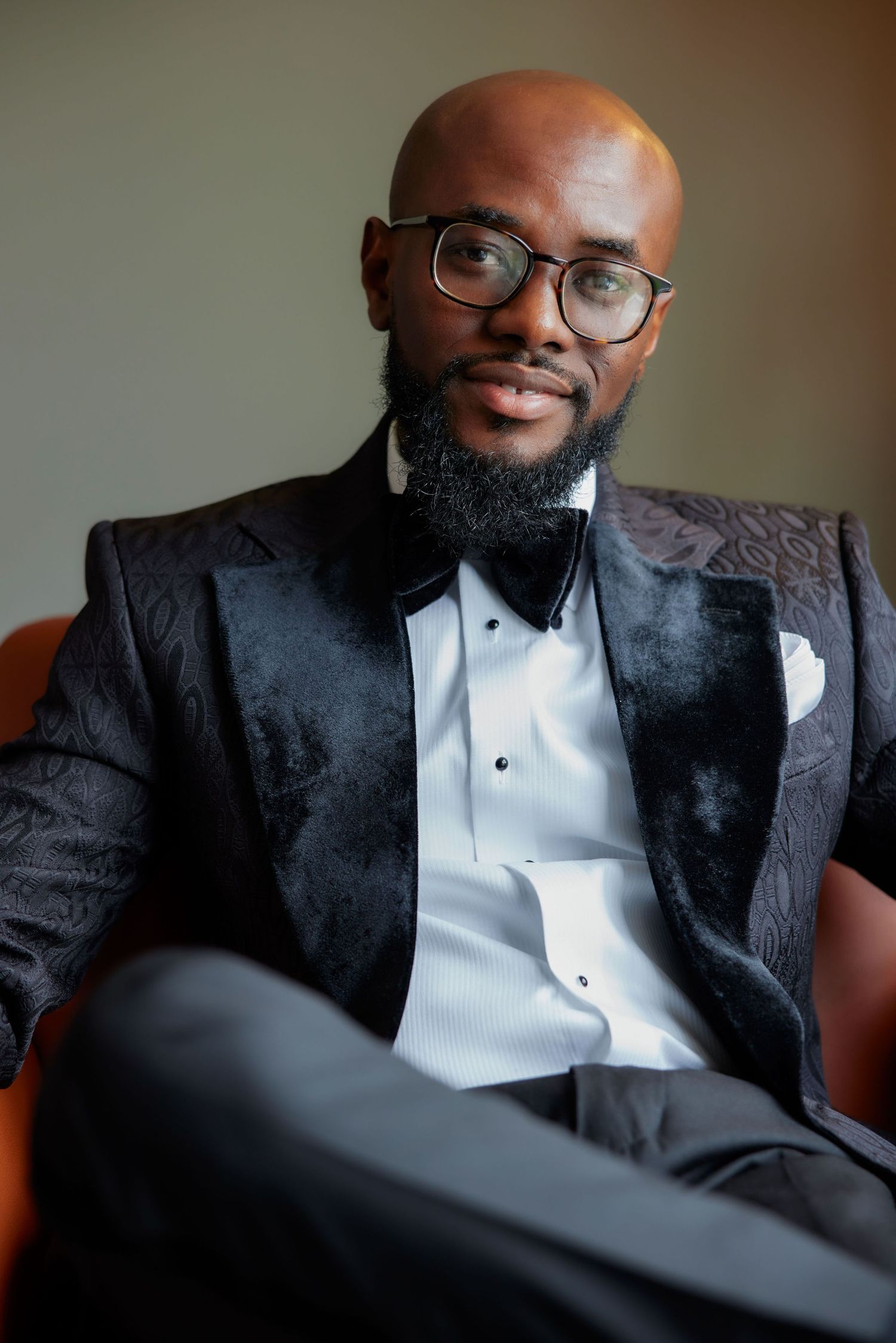 A stylish person in a black patterned suit jacket and white dress shirt sits in a relaxed pose.