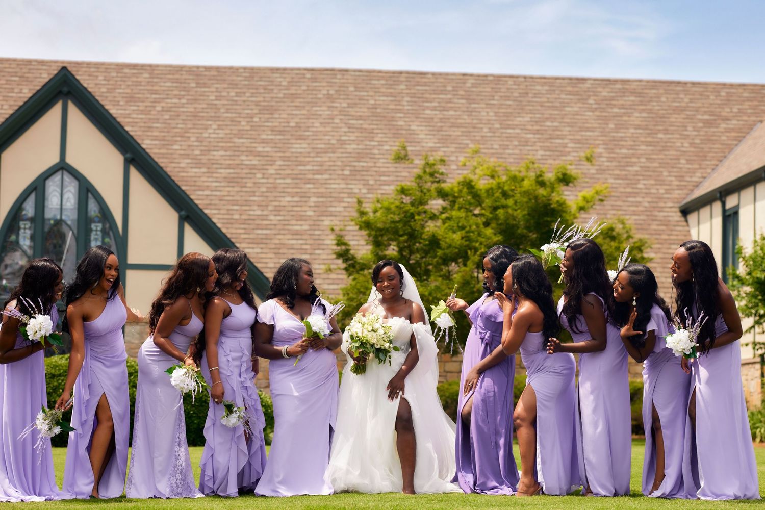 A wedding party in purple bridesmaid dresses and white gowns poses together in a line outdoors.
