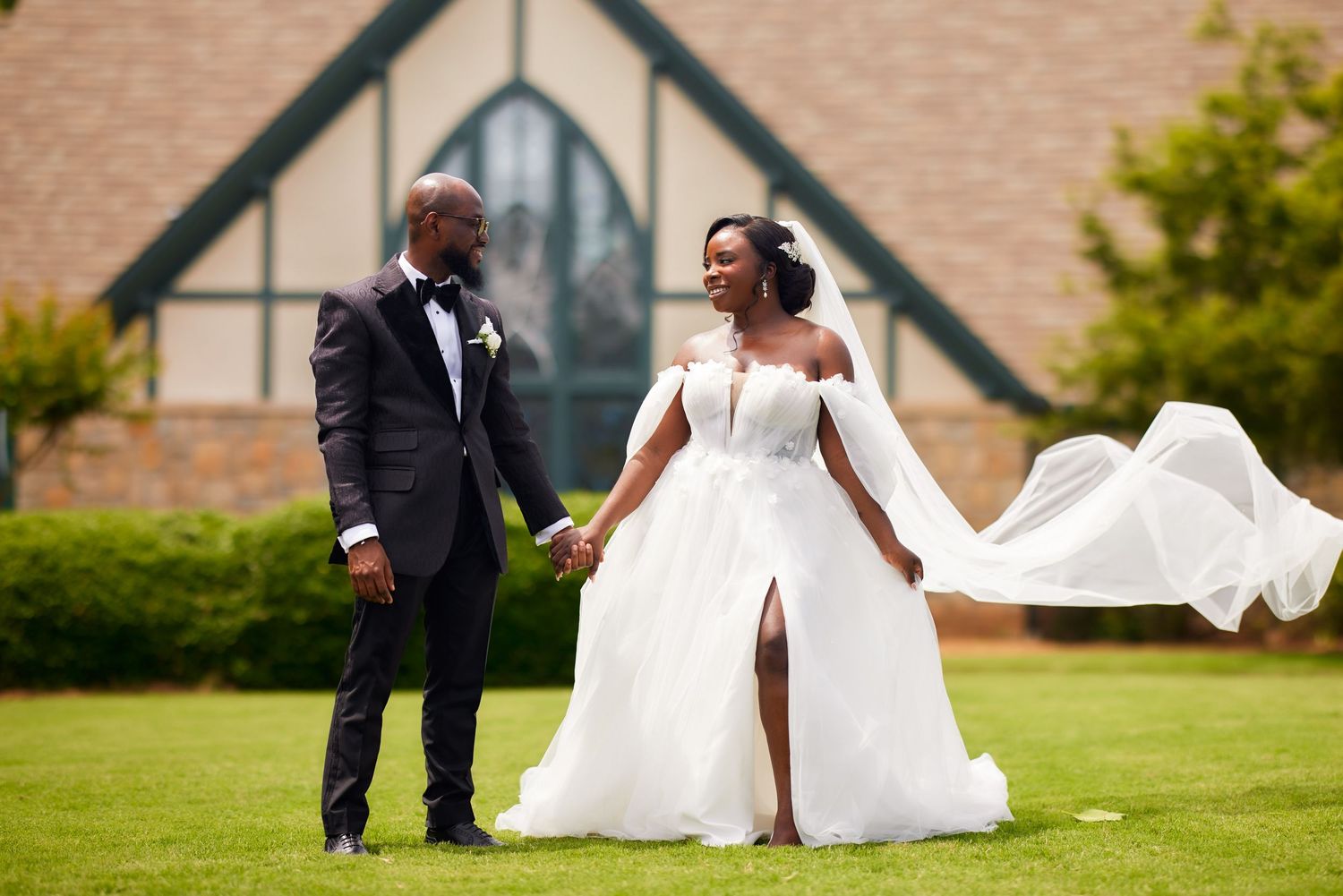 Elegant wedding couple shares a joyful moment together on a green lawn in front of a brick church.
