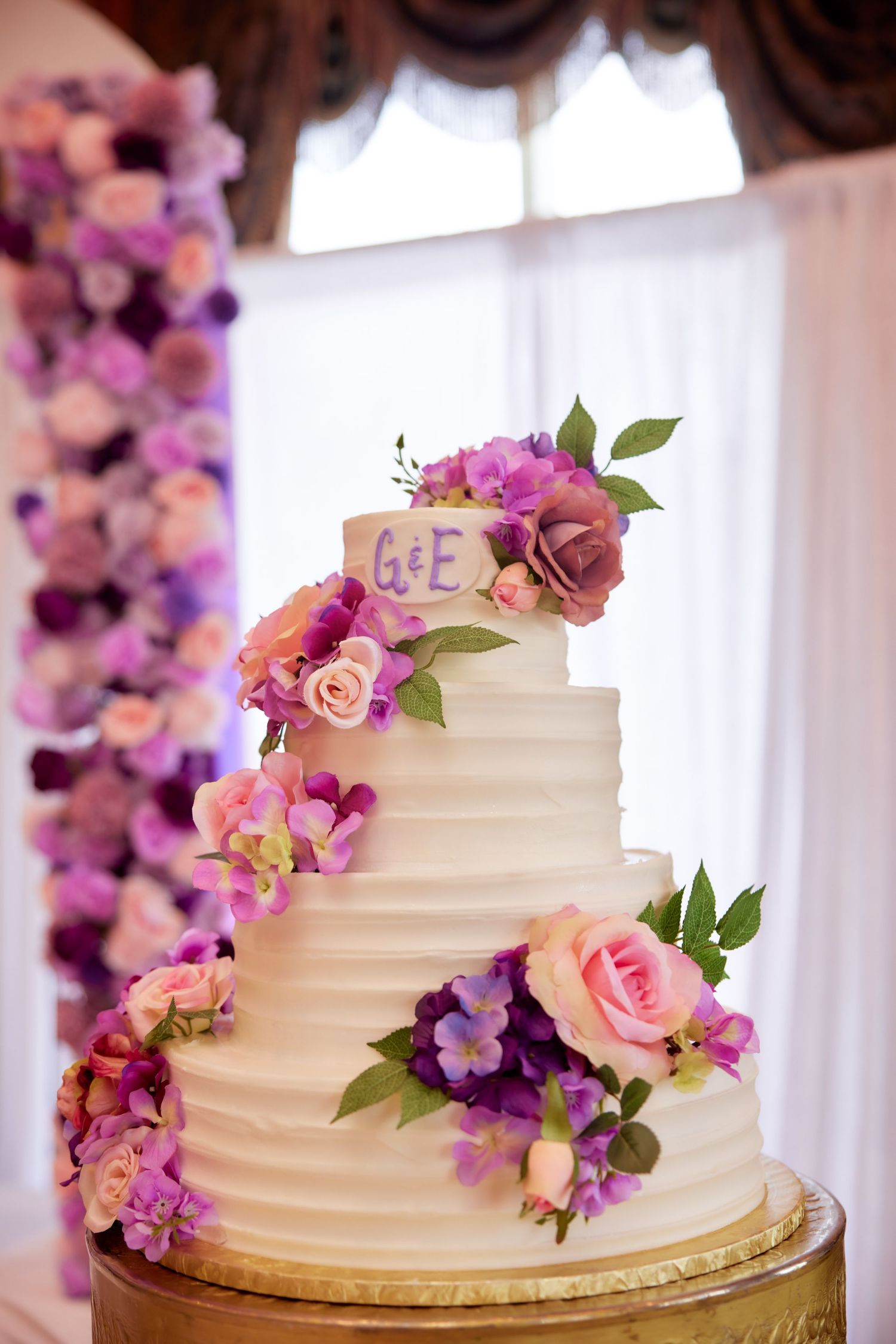A white wedding cake decorated with cascading purple and pink flowers against a bright window backdrop.