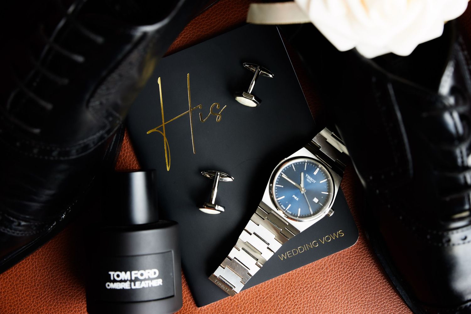 Luxury accessories including Tom Ford perfume, watch, and jewelry elegantly arranged on dark surface with dramatic lighting.