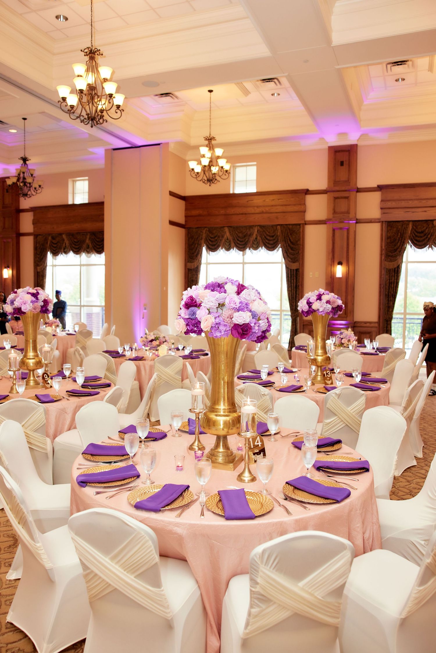 Elegant wedding reception hall with white chair covers, purple napkins, and pink uplighting highlighting ornate chandeliers.