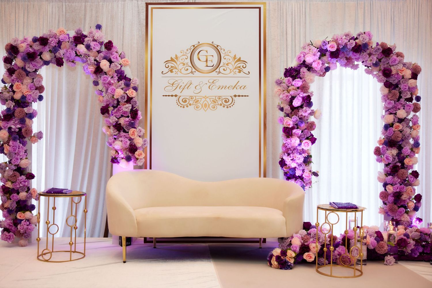 Elegant wedding venue decor featuring gold-framed logo sign and purple balloon arch with floral accents against white backdrop.