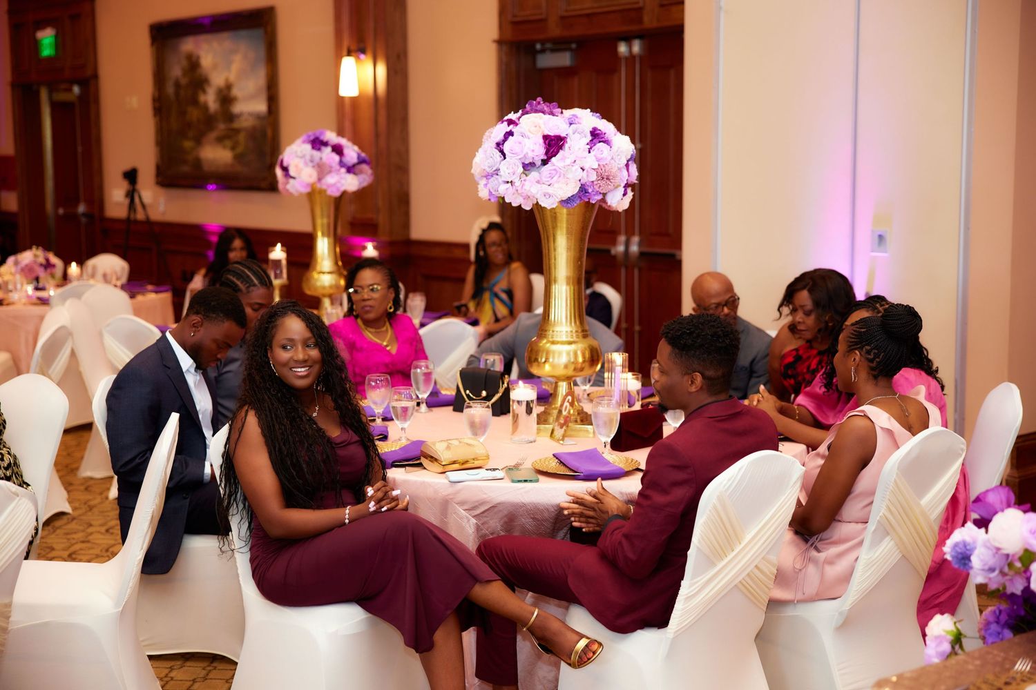 Wedding reception guests dance and celebrate together on the dance floor with pink and purple lighting in a banquet hall.