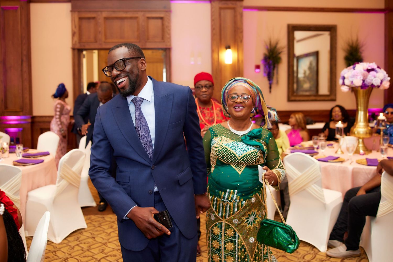 Guests in traditional Nigerian wedding attire celebrate at an elegant reception hall decorated with white linens and pink lighting.