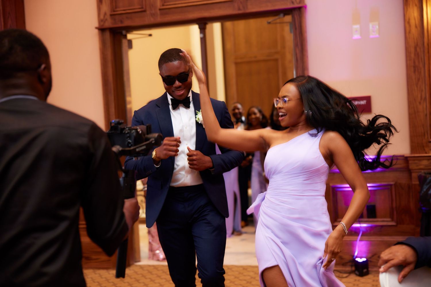 Wedding guests dancing and having fun on the dance floor at an indoor reception venue with purple lighting.