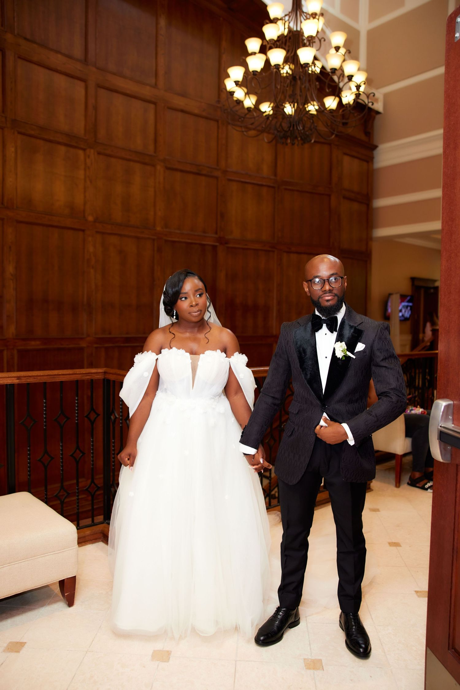 A couple poses together at their elegant wedding, with wood-paneled walls and a crystal chandelier in the background.