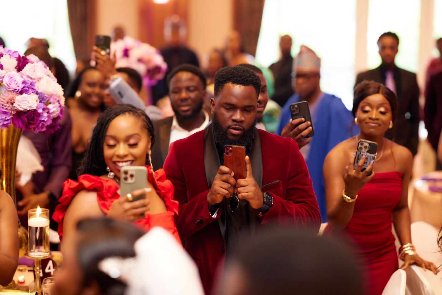 Wedding guests in burgundy attire capture photos and videos with their phones during a celebration.
