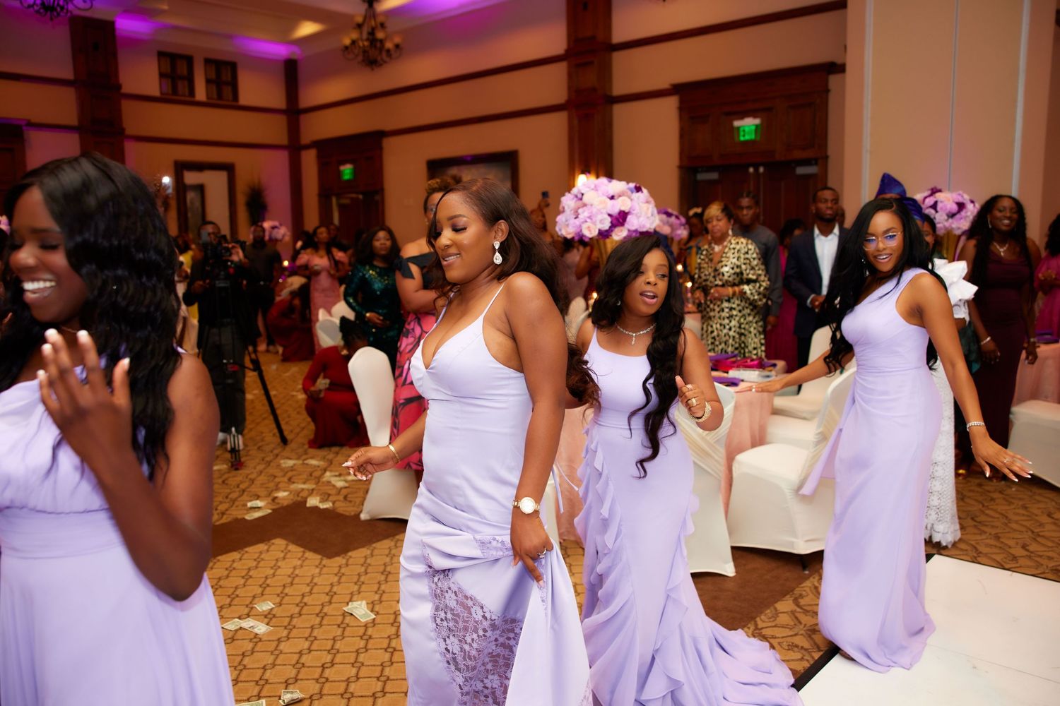 Group of guests in white attire dancing together at a celebration in an elegant ballroom with purple lighting.