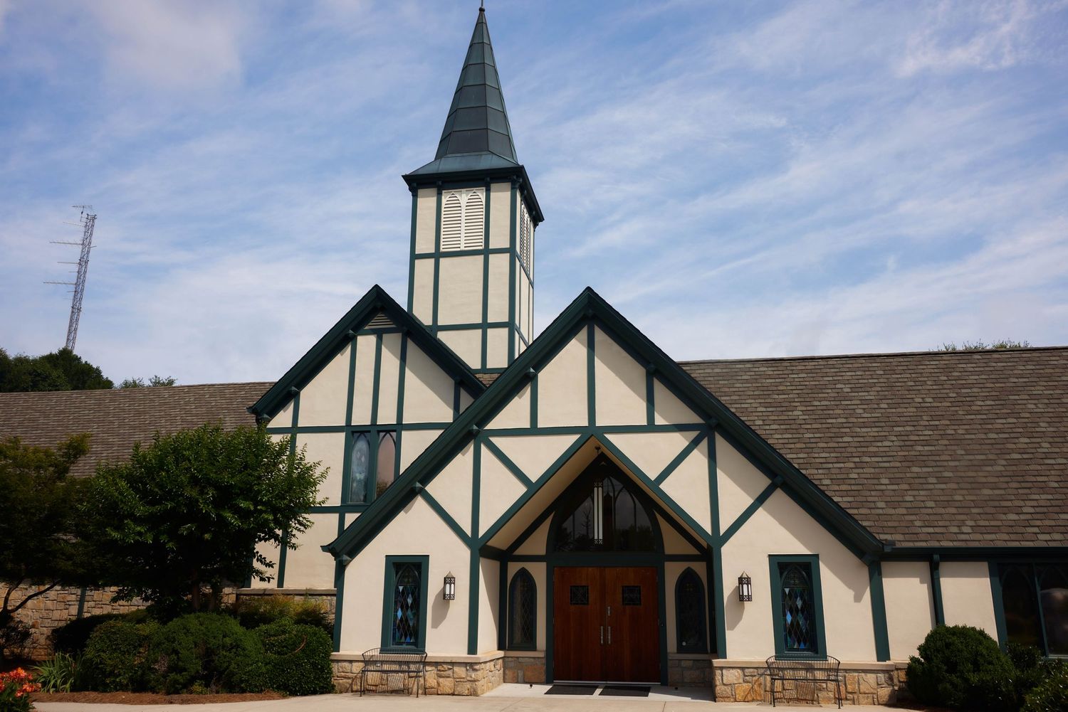 Traditional Tudor-style church building with white walls and dark trim features a tall steeple against a blue sky.