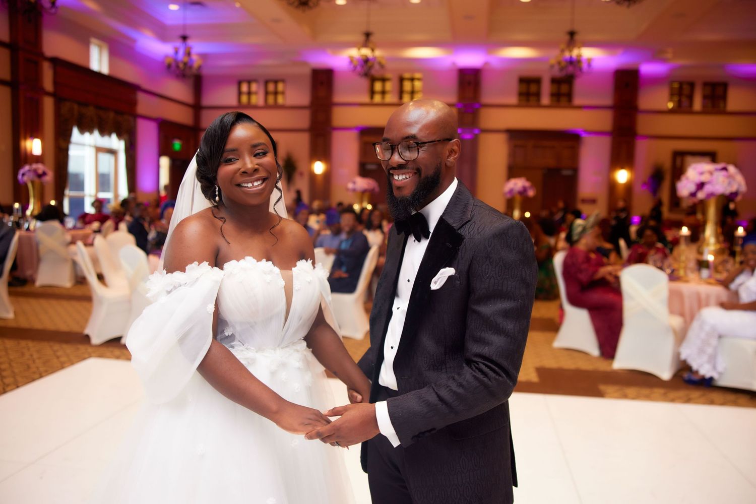 A wedding couple shares a romantic dance moment on an elegantly lit ballroom floor in pink and purple hues.