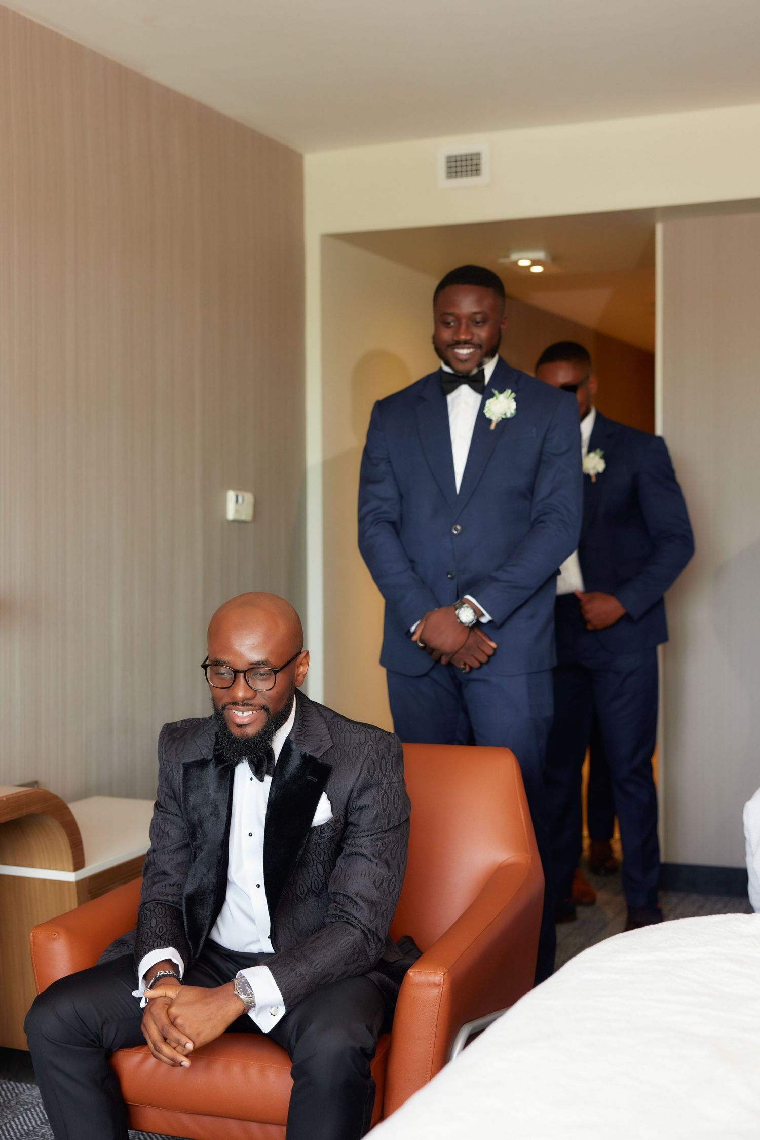 Men in formal navy suits and white boutonnieres sit and stand in a room with tan leather chairs.