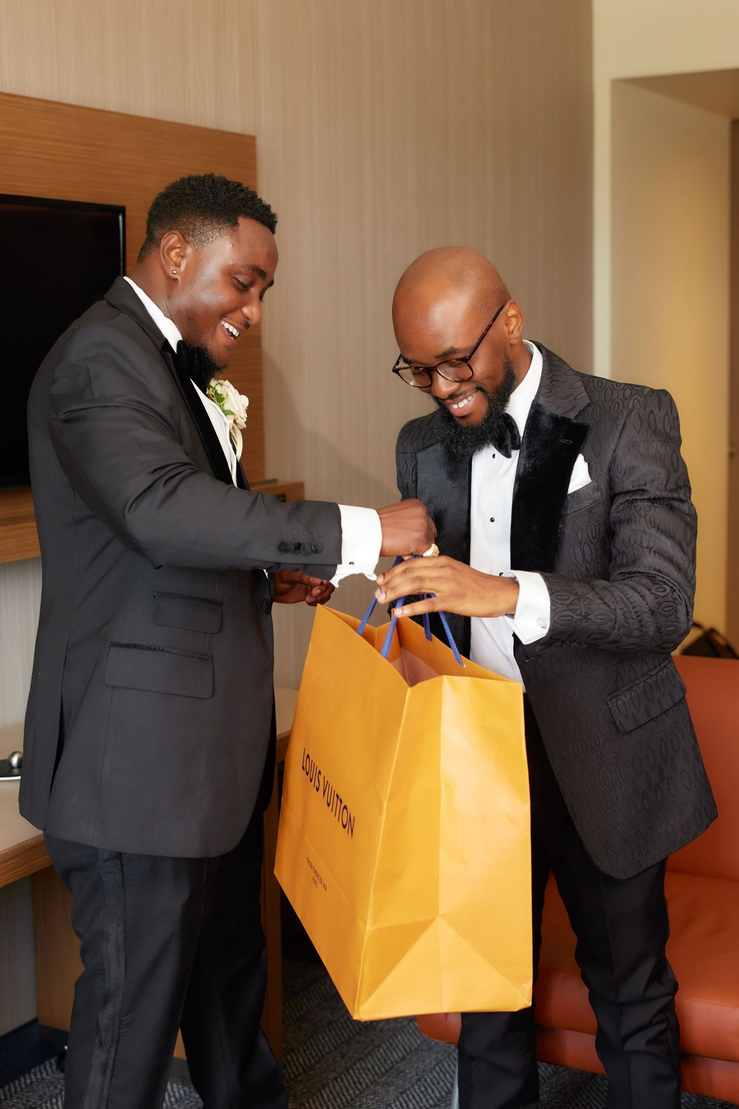 Two men in formal black tuxedos smile while looking inside a luxury shopping bag together.