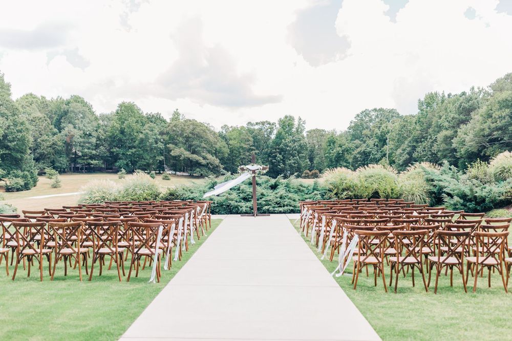 Empty wooden rustic cross back chairs set up in rows on grassy lawn for an outdoor wedding ceremony.