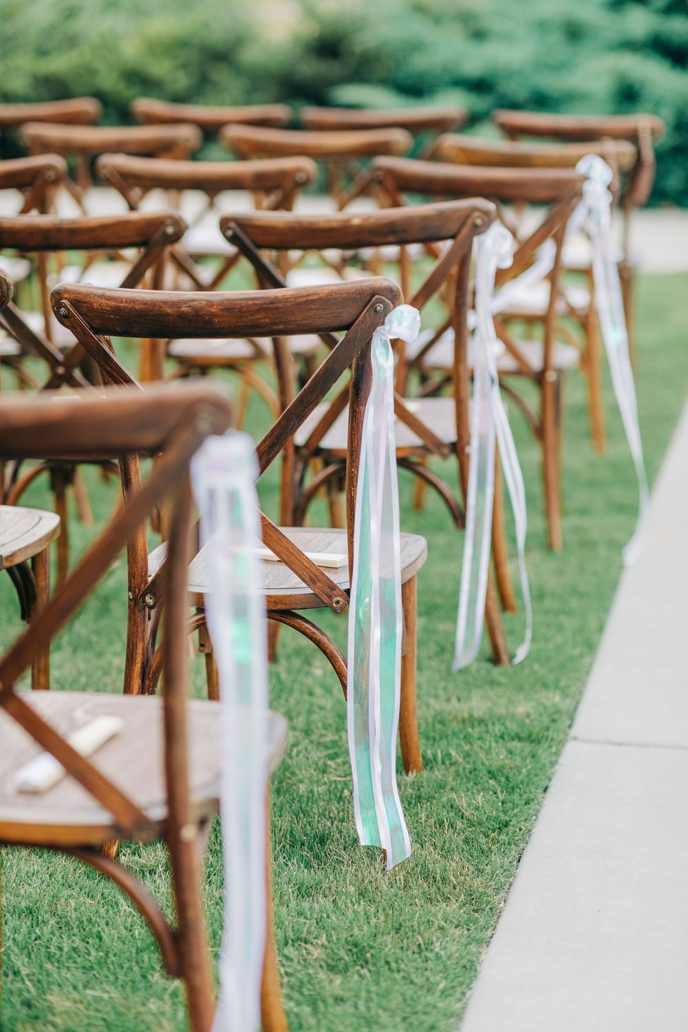 Wooden cross-back chairs with white fabric ribbons line up on a green lawn for an outdoor wedding ceremony.