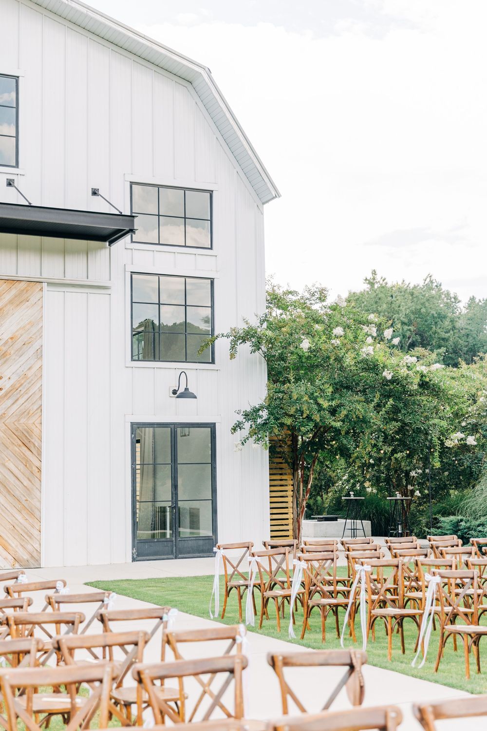 Modern farmhouse wedding venue with white exterior walls and wooden cross-back ceremony chairs set up outdoors at Barn South Wedding Venue