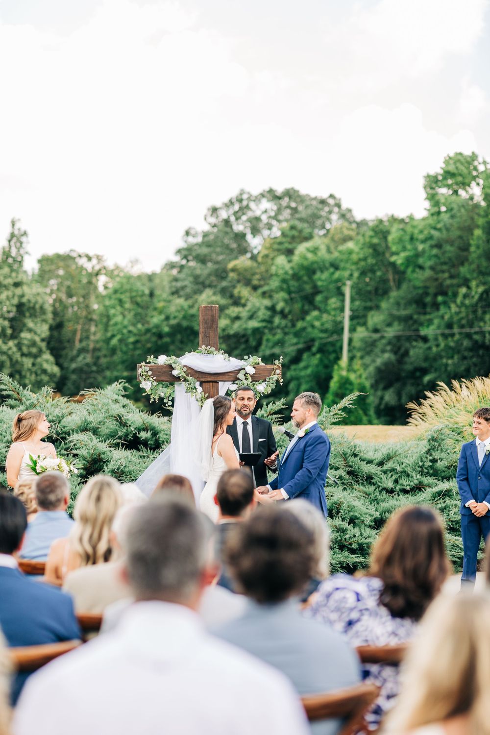 Wide view of outdoor wedding ceremony with guests seated on benches watching couple exchange vows under white arch.