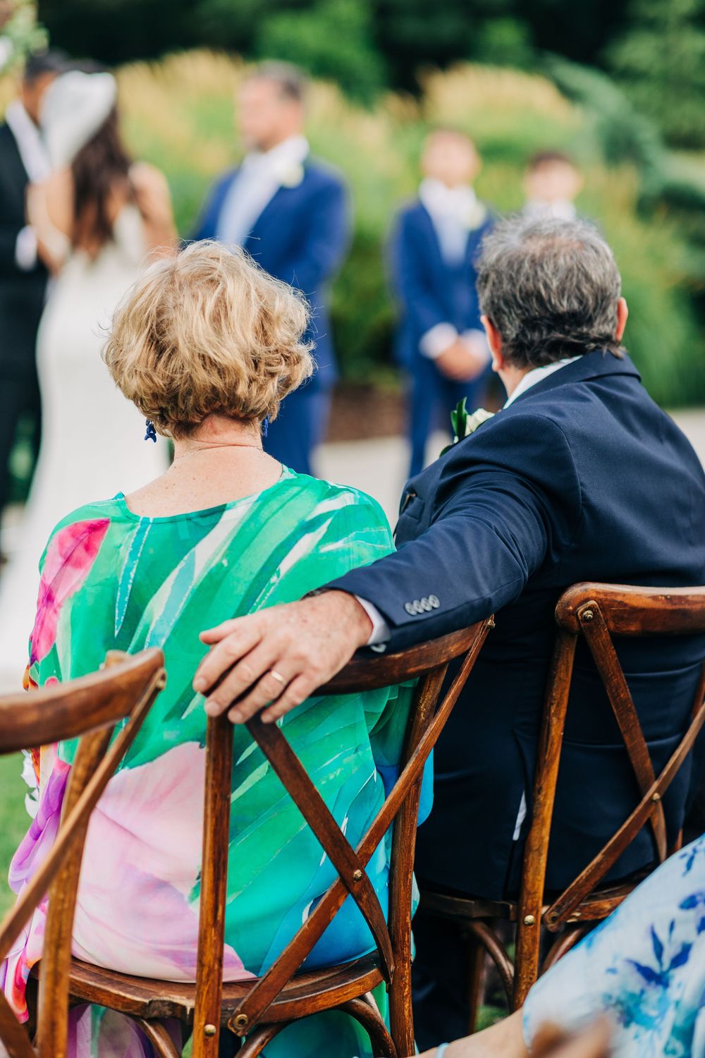 People sit in wooden chairs wearing green and blue attire at an outdoor gathering while socializing.