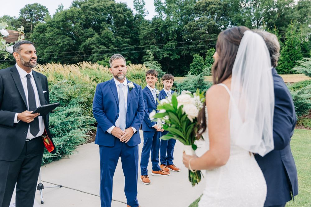 Wedding party on a sunny day with guests wearing formal blue suits and a bride in white outdoors.