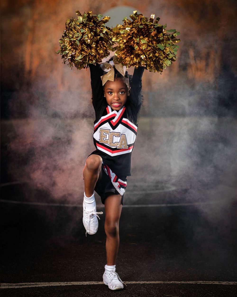 Cheerleader performs high kick with gold pom poms raised against dramatic smoky backdrop.