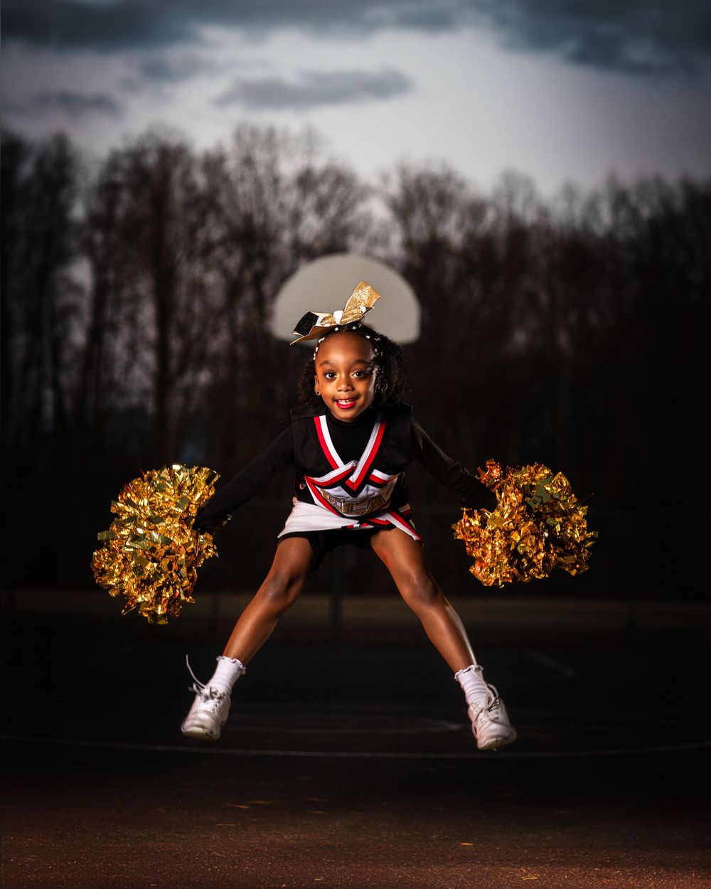 Cheerleader performs dynamic pose with gold pom poms against stormy sky.