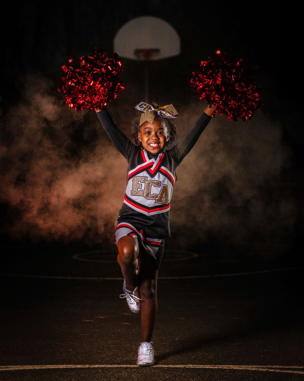 Energetic cheerleader jumps with red pom poms on basketball court at night.