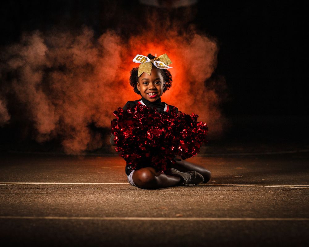 Cheerleader sits with red pom poms against dramatic orange smoke effect.