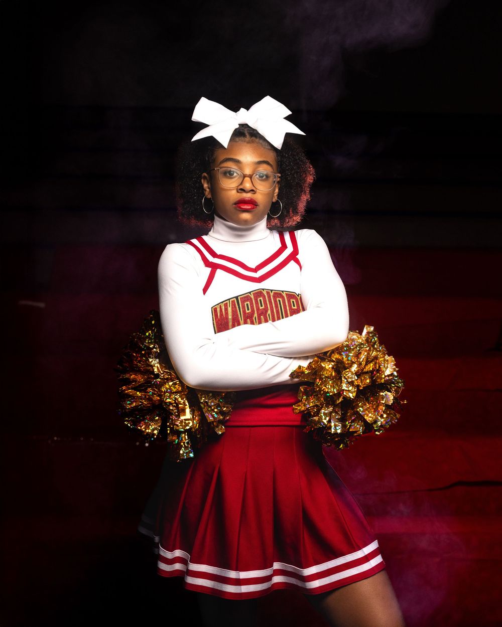 A cheerleader poses in a red and white uniform with gold pom poms against a dark background.