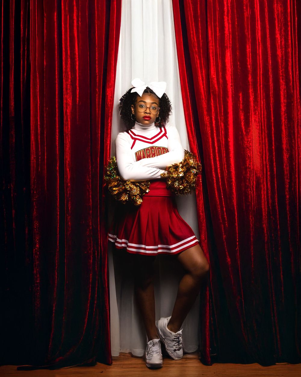 A dramatic portrait of a cheerleader against red curtains in a traditional uniform with pom poms.