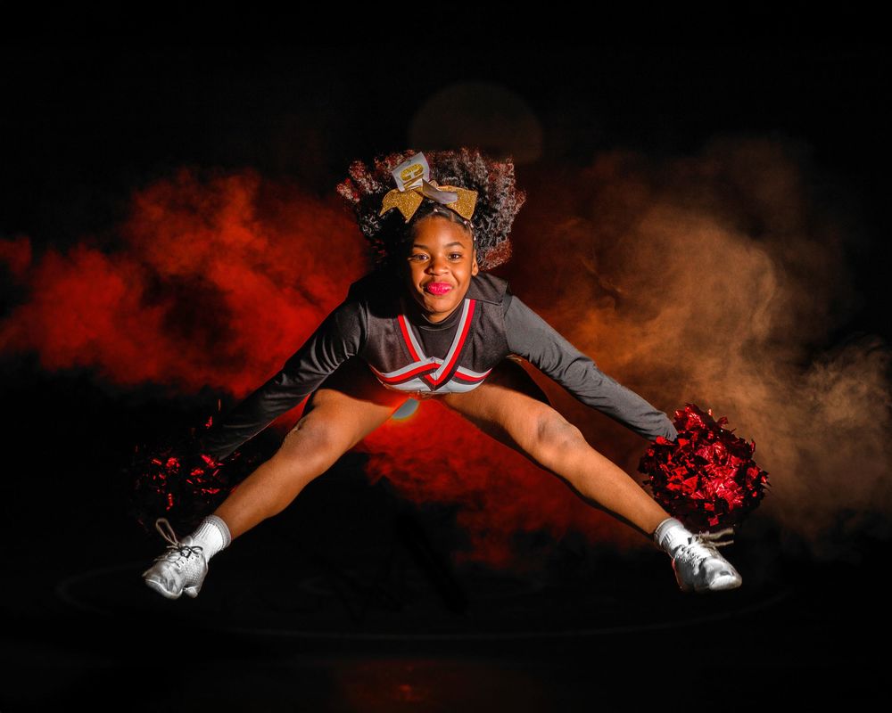 Dynamic cheerleader performs split jump with red pom poms against smoky background.