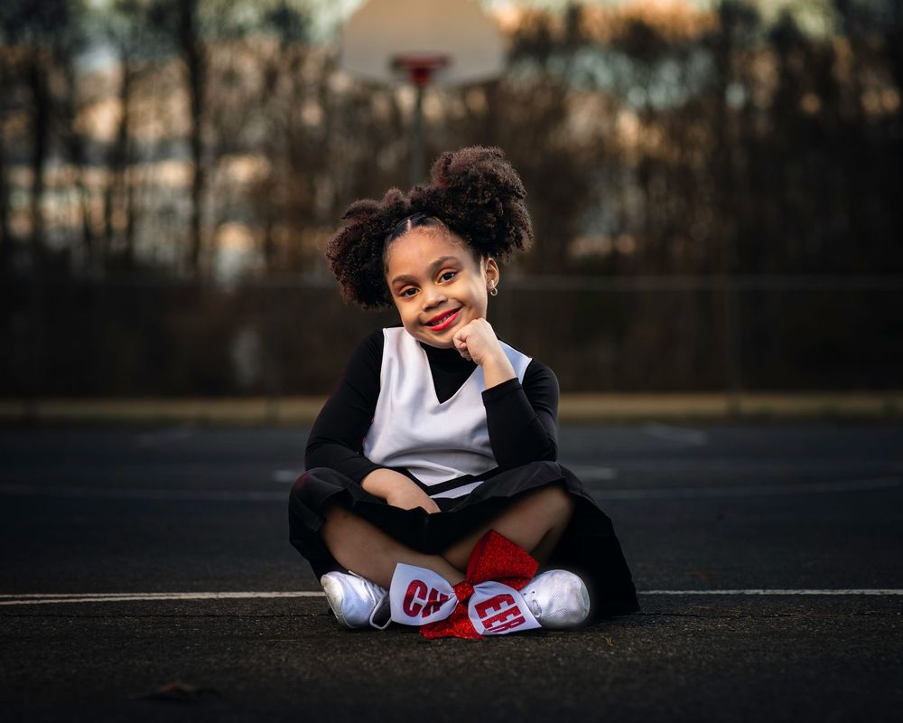 Cheerleader sits casually on outdoor court at sunset wearing team uniform.