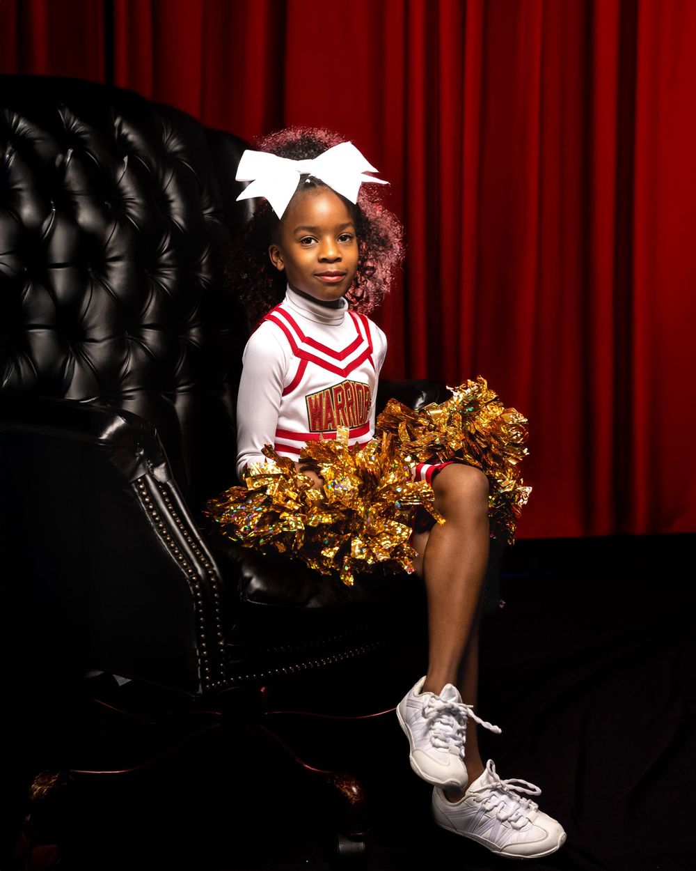 A cheerleader sits elegantly on a black leather couch with gold pom poms and red curtain backdrop.