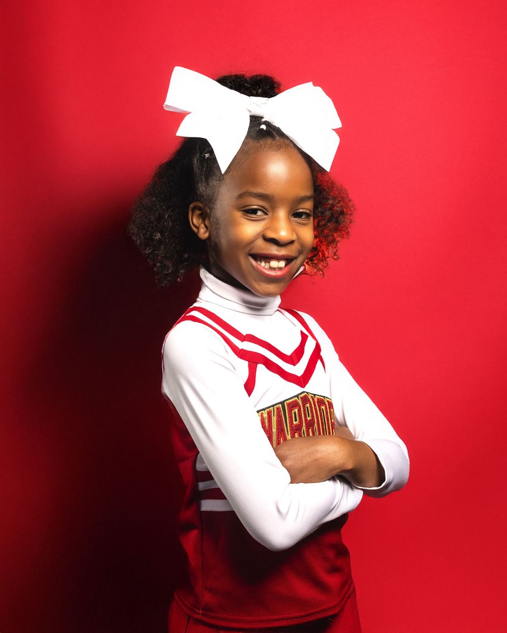 A cheerleader poses confidently against a bright red background in a traditional uniform.