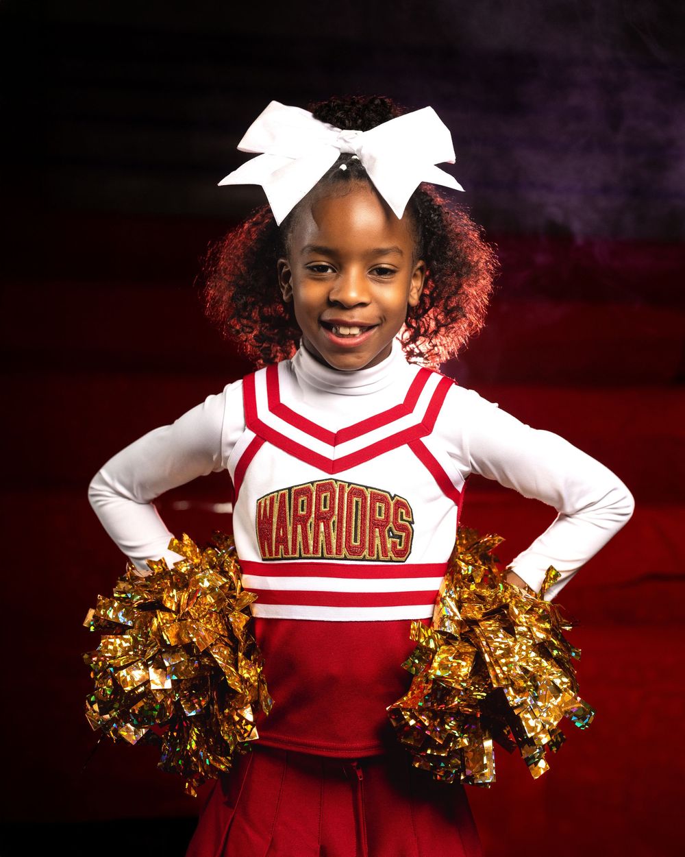 Young cheerleader in red and white Warriors uniform poses with gold pom poms against dark background.