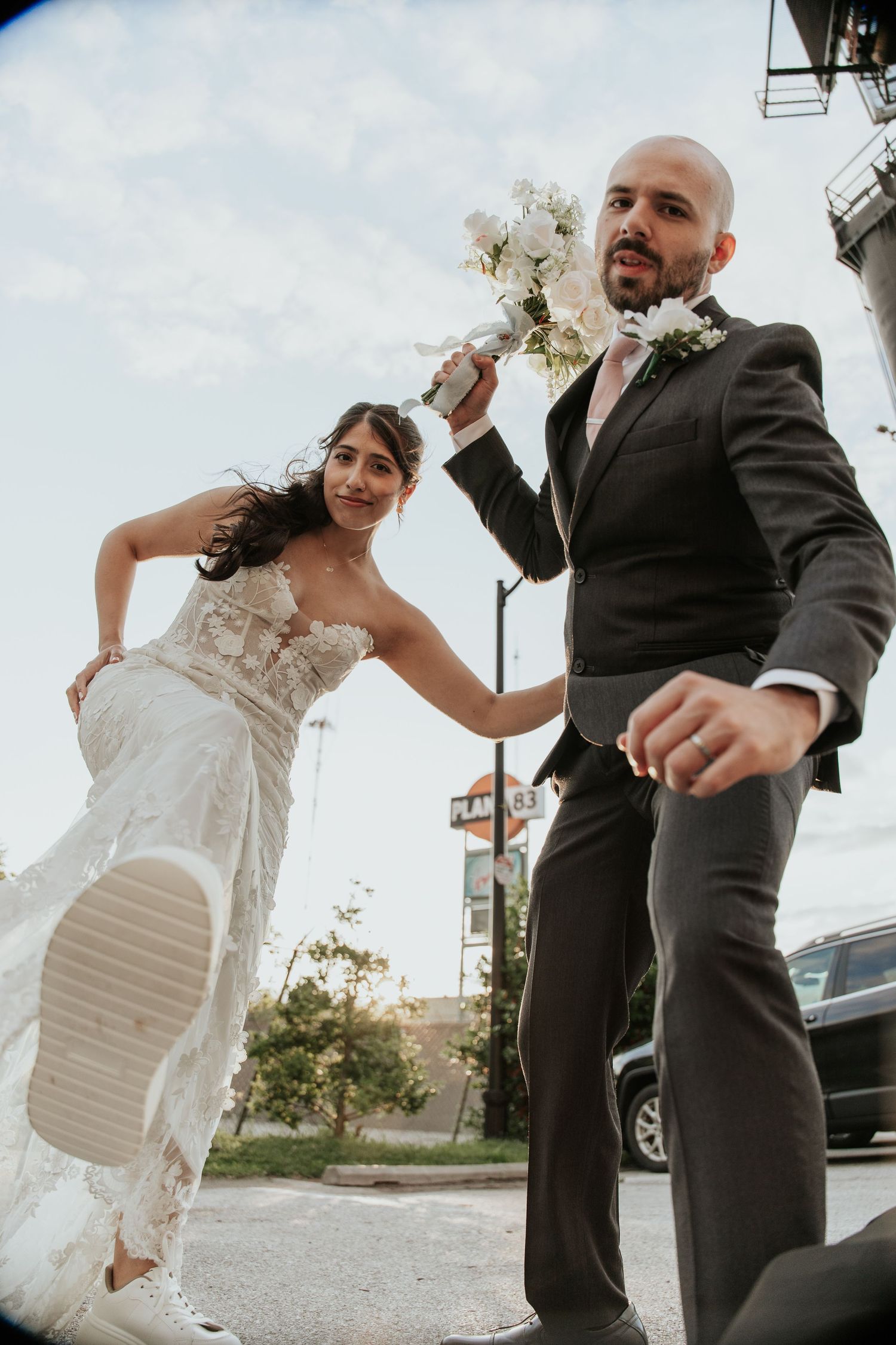 A romantic couple in wedding attire dips in a sunlit meadow at sunset with a bouquet of flowers nearby.