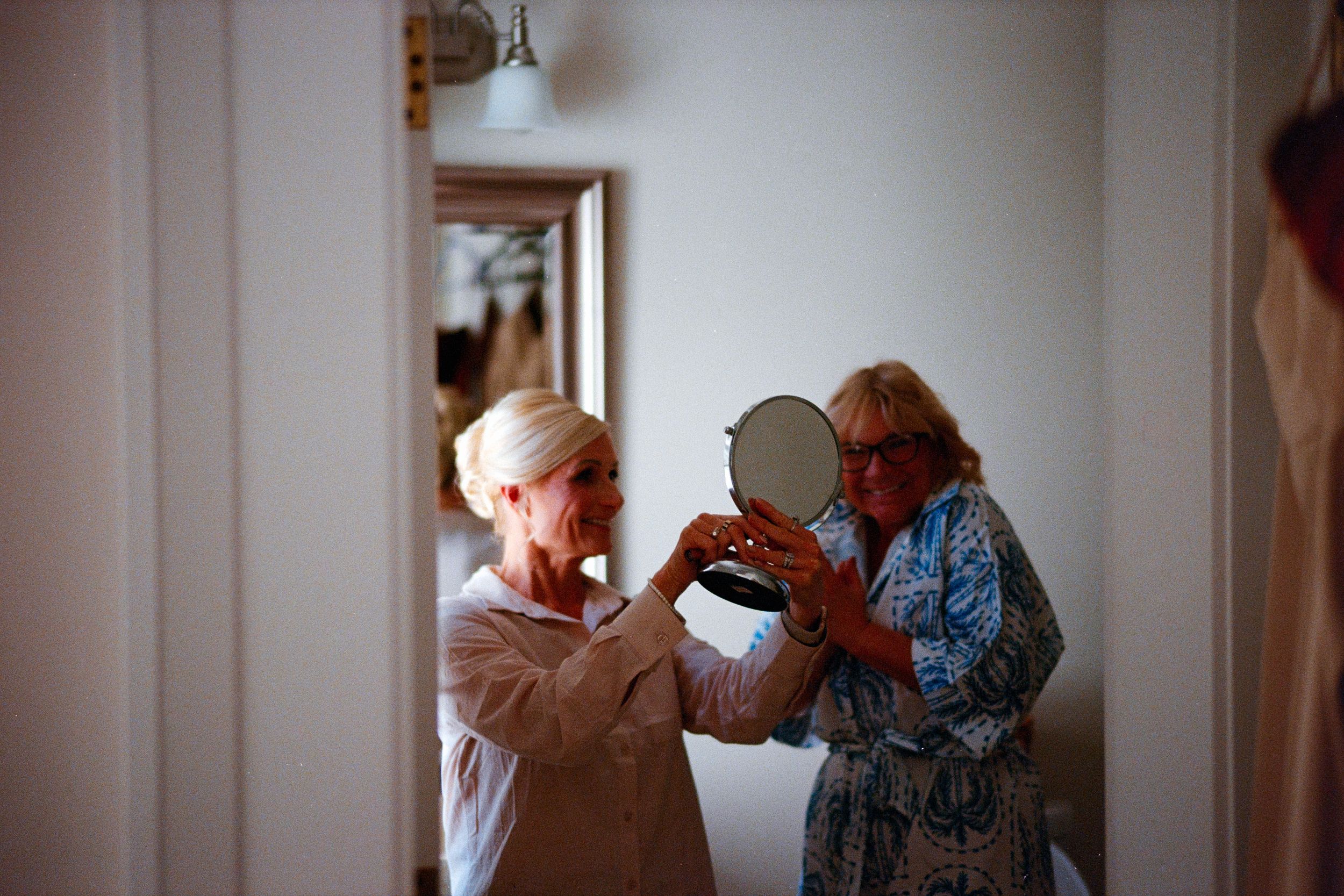 Two elderly people hold up and gaze into a small antique mirror in a white hallway.