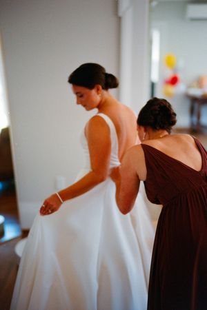 A bride in a white wedding dress being assisted with final preparations by an attendant in a burgundy gown.