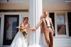 Two people in wedding attire share a joyful moment between white columns on a sunny day.