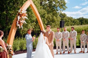 Wedding ceremony outdoors with a wooden arch and groomsmen in light colored suits standing in a line.
