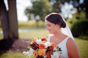Bride holding a vibrant orange and burgundy fall wedding bouquet outdoors on a sunny day.