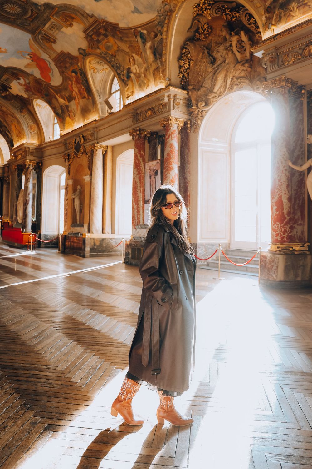 A woman in a long dark trench coat and sunglasses stands in a sunlit ornate baroque palace hall.