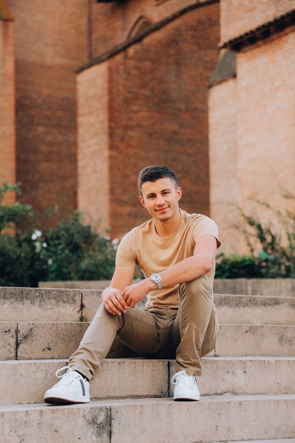 A young man in a beige t-shirt and khaki pants sits relaxed on stone steps near a brick building.