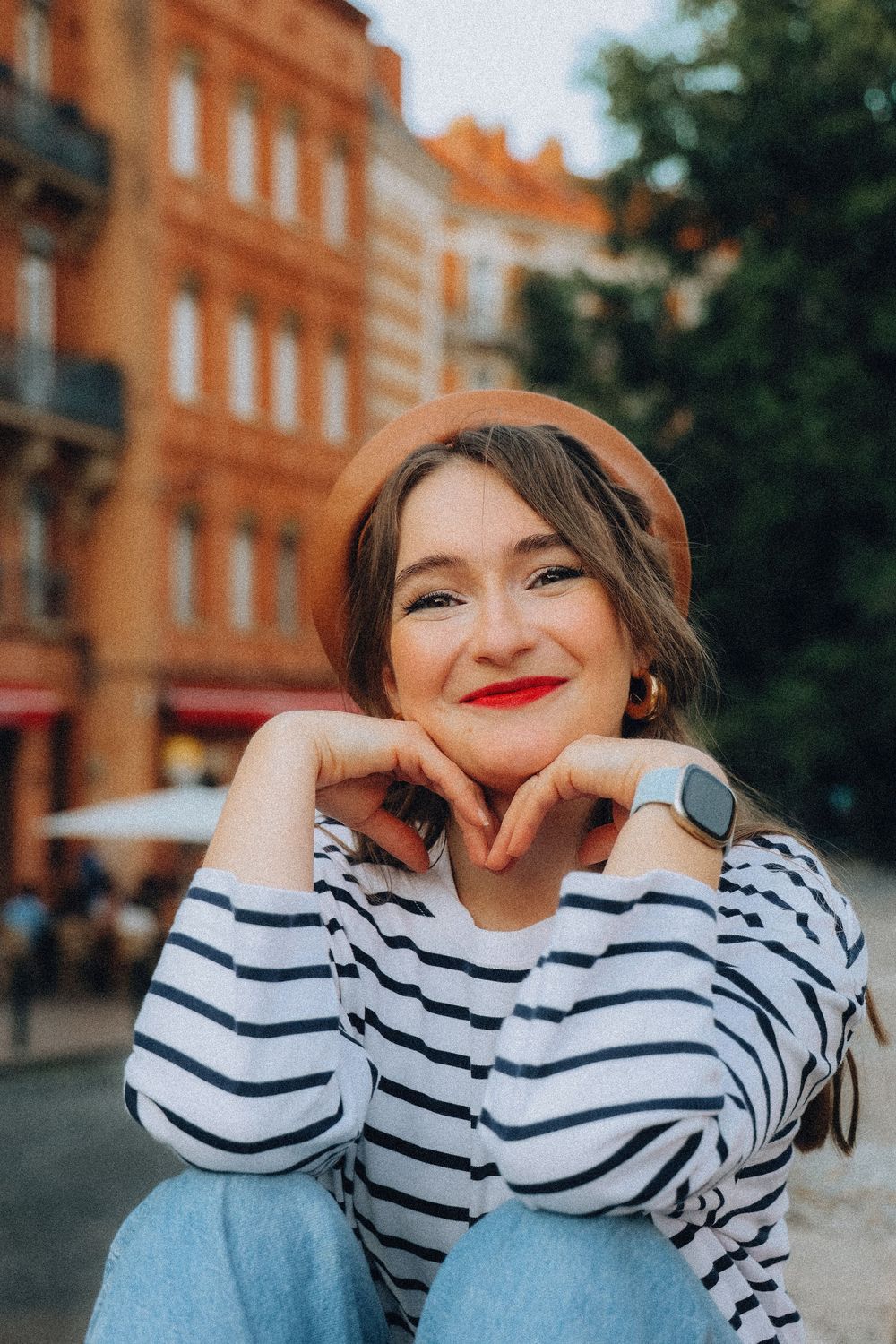 Smiling young woman in striped shirt and brown beret sitting outdoors in urban setting.