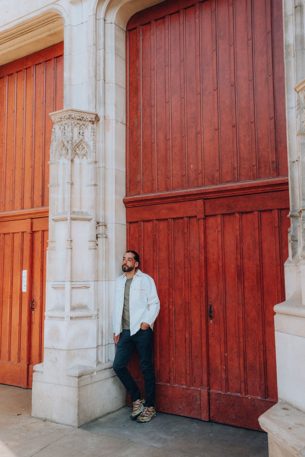 A bearded man in a white jacket leans against ornate stone columns beside large red wooden doors.