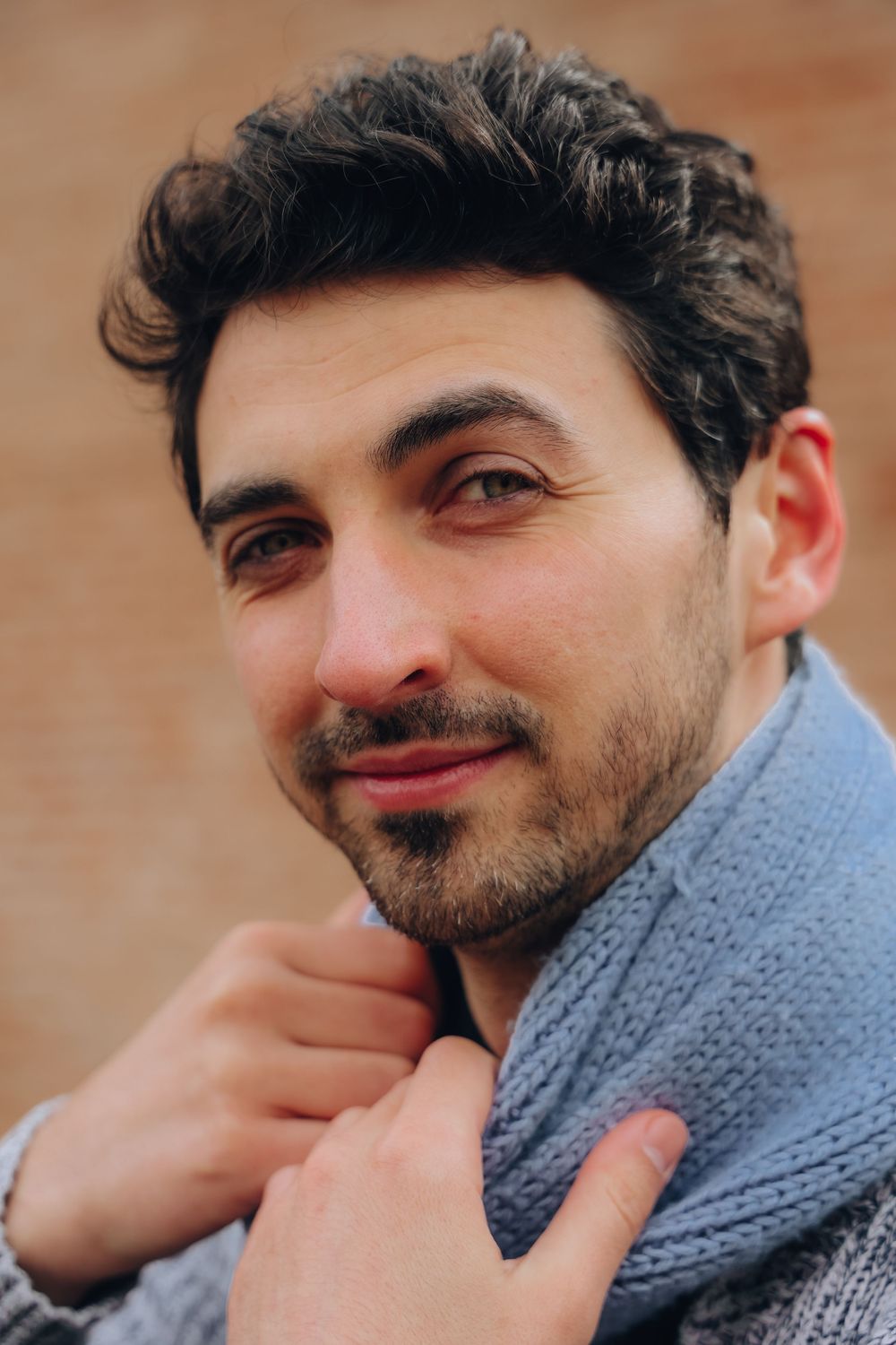 A handsome dark-haired man with light stubble holds his gray knit scarf while smiling warmly outdoors.