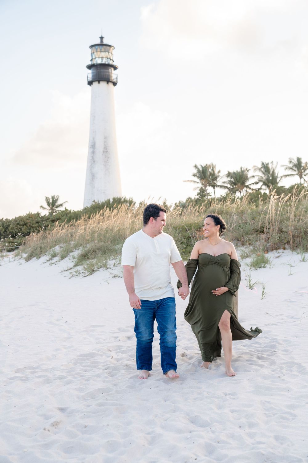 Couple walking along a sandy beach path with a white lighthouse and palm trees in the background.