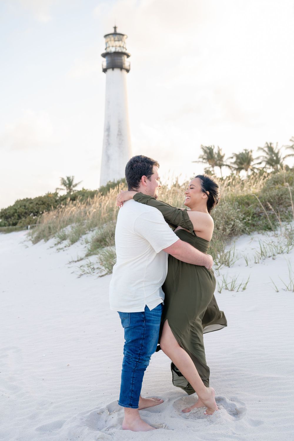 Romantic couple sitting and laughing together on white sand dunes with lighthouse in distance.