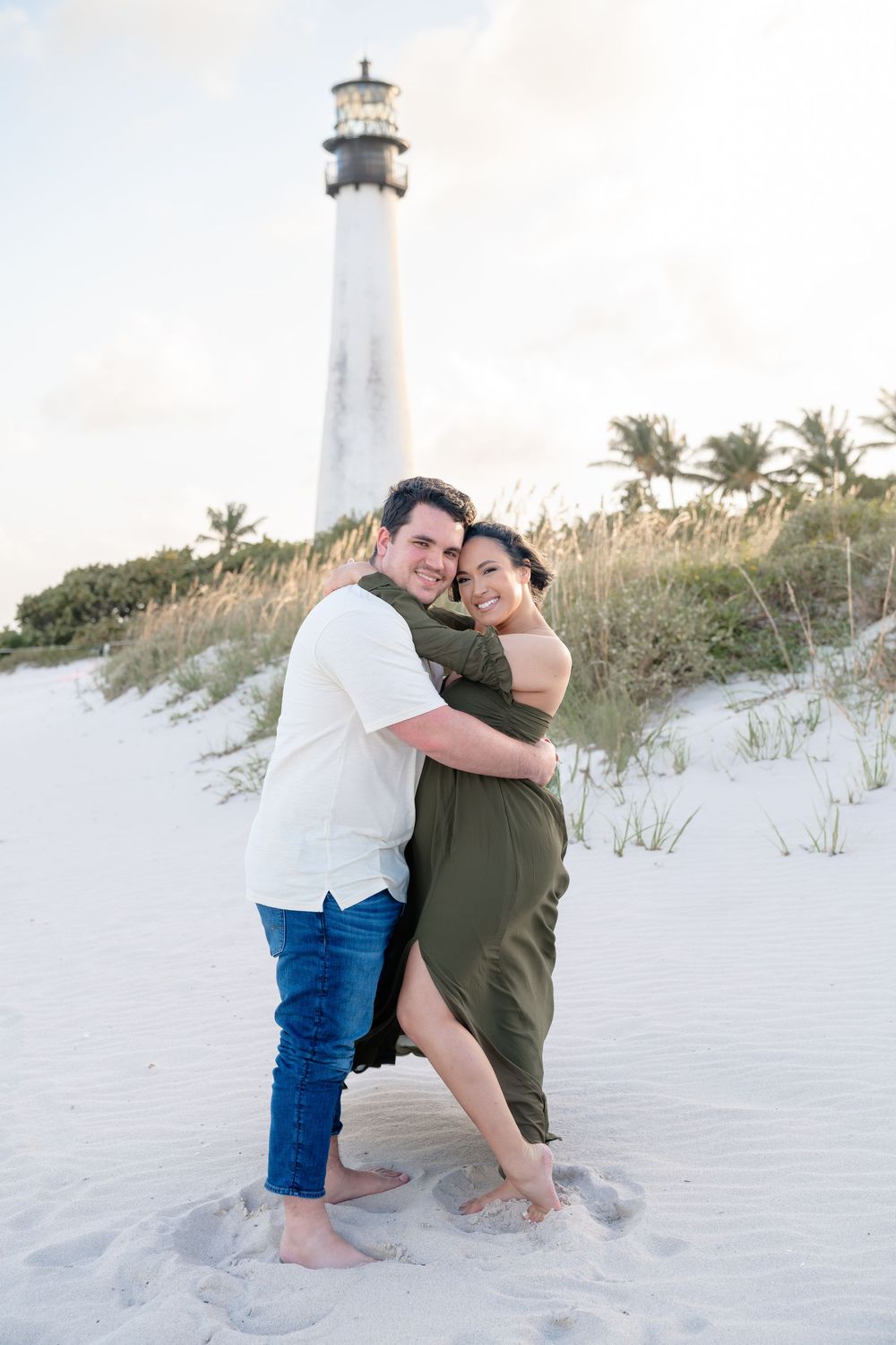 A couple embraces on a white sandy beach near the lighthouse during golden hour.