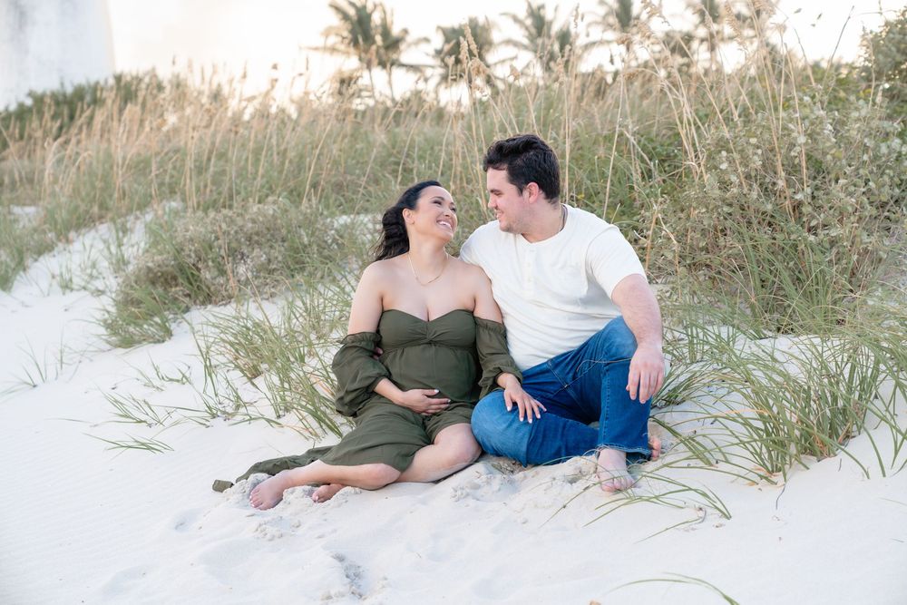 A couple sits together intimately on white sandy dunes with palm trees in the background at sunset.